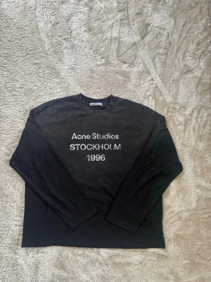 Acne Studios - Sleeve - Svart långärmad tröja från Acne Studios med tryck i vitt på bröstet där det står 'Acne Studios STOCKHOLM 1996'. Tröjan har rund halsringning och avslappnad passform, perfekt för en stilren och enkel look.