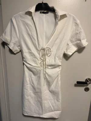 Vit kortklänning med snörning - Kort vit klänning från Zara med krage, korta ärmar och snygg snörning framtill i midjan. Klänningen har en v-ringad öppning och är gjord i ett lätt material som passar perfekt till varma dagar.