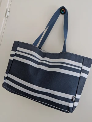 Blåvit randig tygväska strandväska - Snygg blå strandväska med vita horisontella ränder och dubbla axelband. Väskan är rymlig och har en enkel, stilren design som passar perfekt till shopping eller stranden. Tillverkad i slitstarkt tygmaterial. Jättefint skick.