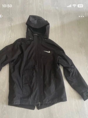 Svart regnjacka från Nike med huva - Snygg svart regnjacka från Nike med vit logga på bröstet. Jackan har huva, dragkedja framtill och justerbara ärmslut. Tillverkad i vattenavvisande material, perfekt för regniga dagar. Enkel och stilren design som passar till det mesta.