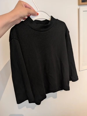 Svart ribbad kort topp - Säljer en svart ribbad topp med kort ärm och hög hals. Toppen har en croppad passform och är tillverkad i ett mjukt, stretchigt material som sitter snyggt på kroppen. Perfekt att styla med jeans eller kjol för en clean look.