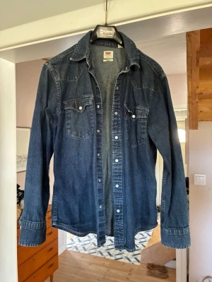 Mörkblå jeansjacka från Levi's - Klassisk mörkblå jeansjacka från Levi's med tryckknappar framtill och två bröstfickor med lock. Säljer då den är för liten