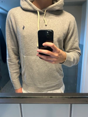 Grå hoodie från Polo Ralph Lauren - Snygg grå hoodie från Polo Ralph Lauren med klassisk broderad logga på bröstet. Hoodien har huva med vita dragsnören och en stor magficka framtill. Tillverkad i mjukt sweatshirtmaterial och har ribbade muddar vid ärmar och nederkant.