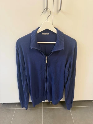 Marinblå ziptröja från Gran Sasso, stl 50/M - Snygg marinblå ziptröja från Gran Sasso med ribbad krage och dragkedja framtill. Tillverkad i Italien och har en stilren, enkel design som passar perfekt till jeans eller chinos. Materialet känns mjukt och lätt, perfekt för lager på lager.