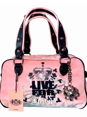 Rosa Juicy Couture handväska - Söt rosa handväska från Juicy Couture med svarta detaljer, silverfärgad krona och texten 'LIVE FOR Juicy' framtill. Väskan har dubbla handtag med nitar, hjärtformad detalj med J på baksidan och en stor metallberlock. Perfekt för dig som gillar statement-accessoarer.