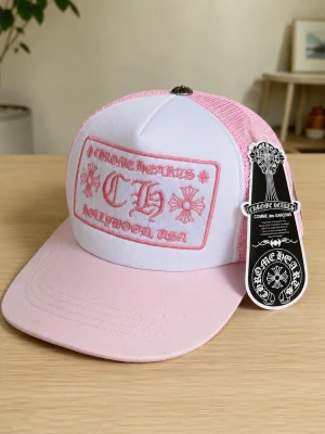 Chrome Hearts keps - Kepsen är ljusrosa och vit med broderad logga och text på framsidan. Klassisk trucker-modell med mesh på sidorna och justerbar snapback. Perfekt för dig som gillar streetwear och unika accessoarer.