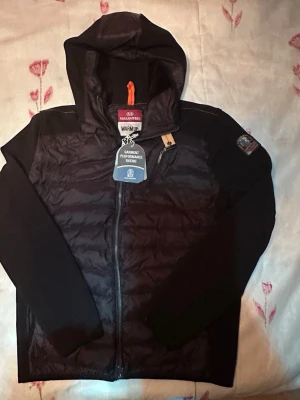 Svart parajumper cardigan oanvänd helt ny med huva - Snygg svart pufferjacka med huva och dragkedja framtill. Jackan har quiltad front och rygg, samt släta ärmar. Märkespatch på ena ärmen och praktisk bröstficka med dragkedja. Perfekt för kalla vinterdagar och riktigt varm. Den är i storlek L då den är i liten storlek för och vara XL så den är L…