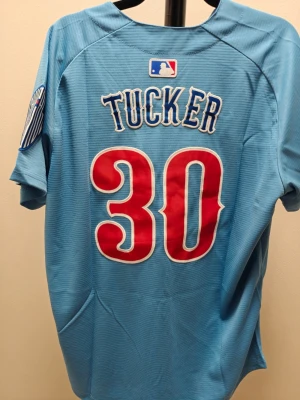 Blå Chicago Cubs baseballtröja - Snygg ljusblå baseballtröja från Nike med Chicago Cubs-logga och nummer 30 i rött och vitt. Namnet 'Tucker' står på ryggen. Tröjan har korta ärmar, knappar framtill och patch på ärmen. Perfekt för sport eller streetwear.