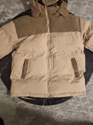 Beige pufferjacka med manchesterdetaljer - Säljer en beige pufferjacka med bruna manchesterdetaljer på axlar, huva och fickor. Jackan har dragkedja framtill, två fickor och en mysig huva. Perfekt för kalla dagar och riktigt skön att bära.