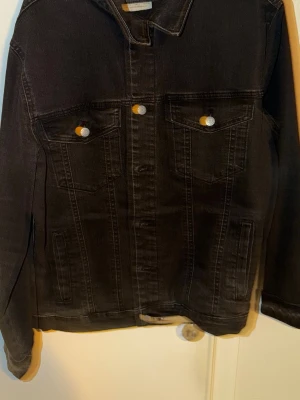 Svart jeansjacka med silverknappar - Stilren svart jeansjacka med klassisk passform och silverfärgade knappar. Jackan har två bröstfickor med lock och knapp samt två sidofickor. Perfekt för dig som gillar en tidlös look och vill ha en jacka som funkar till det mesta.