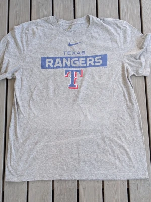 Grå Texas Rangers t-shirt från Nike - Snygg grå t-shirt från Nike med Texas Rangers-tryck i blått och rött framtill. Klassisk rund hals och korta ärmar. Perfekt för dig som gillar sportiga och avslappnade plagg. Tillverkad i mjuk bomull för skön känsla hela dagen.