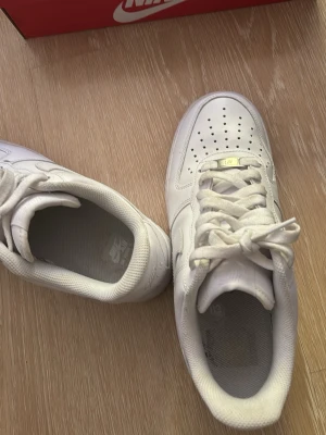 Nike Air Force 1 vita sneakers - Klassiska vita Nike Air Force 1 sneakers i lågt utförande. Skorna är tillverkade i slätt läder med perforerad tåbox för ventilation och har den ikoniska swoosh-loggan på sidan. Tjock sula med AIR-dämpning och vita skosnören. Perfekta för en clean och stilren look.