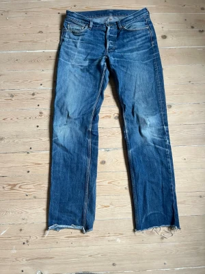 Blå raka jeans med rå kant - Säljer ett par blå jeans med rak passform och slitna detaljer. Jeansen har råa, fransiga benslut och klassiska fem fickor. Materialet är denim i bomull och färgen är tvättad blå med ljusare partier och slitningar för en vintage look. Byxorna har en inre lagning på rumpan och höger innerficka är nylagad, visas på bilderna. Vänster ficka har hål i sig men går enkelt att laga. Priset är ej skrivet i sten!