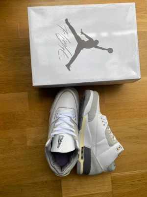 Vita och gråa Air Jordan 3 sneakers - Snygga Air Jordan 3 sneakers i vitt och ljusgrått med detaljer i beige och guld. Skorna har en klassisk siluett med lågt skaft, ovandel i läder och mocka samt synlig Air-enhet i sulan. Perfekta för dig som gillar streetwear och ikoniska sneakers.