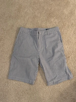 Randiga shorts från Polo Ralph Lauren - Säljer ett par klassiska blå- och vitrandiga shorts från Polo Ralph Lauren. De har bälteshällor, knapp och dragkedja framtill samt en relaxed passform. Perfekta för varma dagar och tillverkade i mjuk bomull för extra komfort.