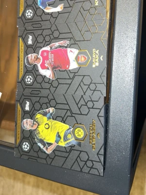 Fotbollssamlarbilder Topps UCL - Två samlarbilder från Topps med fotbollsspelarna Bukayo Saka (Arsenal) och Giovanni Reyna (Borussia Dortmund). Korten har svart bakgrund med guld- och klubbdetaljer samt spelarnas statistik på baksidan. Perfekt för dig som gillar fotboll och samlarprylar.