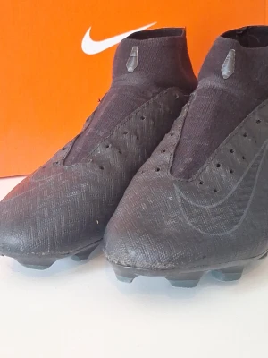 Nike Mercurial svarta fotbollsskor - Säljer ett par svarta Nike Mercurial fotbollsskor med hög ankel och strumpliknande passform. Skorna har diskret Nike-logga, mönstrad ovandel och fasta dobbar för spel på gräs. Tillverkade i syntetmaterial för bra grepp och kontroll på planen.