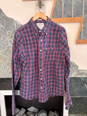 Rutig skjorta från Abercrombie & Fitch - Säljer en klassisk rutig skjorta från Abercrombie & Fitch i blått och rött. Skjortan har lång ärm, knappstängning framtill och en bröstficka. Perfekt för dig som gillar en avslappnad men ändå stilren look.