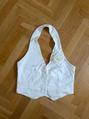 Vit halterneck väst med blomdetalj - Stilren vit väst i halterneck-modell med tre knappar framtill och dekorativ tygblomma på ena sidan. Västen har figurnära passform och två dekorfickor framtill. Perfekt för dig som vill sticka ut med en unik detalj.