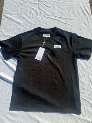 Svart t-shirt Maison Margiela - Svart t-shirt från Maison Margiela med klassisk rund hals och korta ärmar. T-shirten har den ikoniska Margiela-etiketten framtill och är tillverkad i mjuk bomull. Enkel och stilren design som passar till allt.