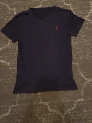 Mörkblå t-shirt från Polo Ralph Lauren - Snygg mörkblå t-shirt från Polo Ralph Lauren med klassisk röd broderad logga på bröstet. T-shirten har korta ärmar och är gjord i mjuk bomull för en skön känsla. Perfekt till jeans eller shorts för en clean och enkel stil.
