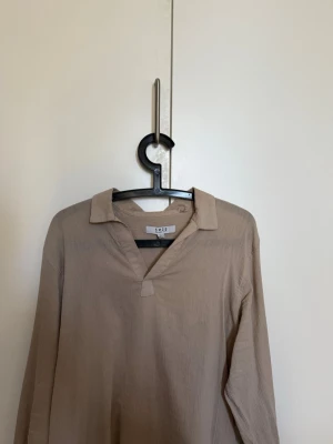 Beige tröja - Stilren beige tröja med krage och v-ringad hals. Tröjan har lång ärm och är tillverkad i ett mjukt, lätt material som ger en avslappnad känsla. Perfekt för dig som gillar minimalistisk och enkel stil.