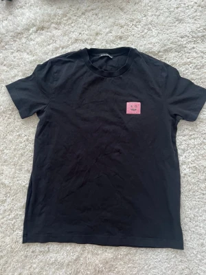 Svart Acne Studios t-shirt med patch - Säljer en svart t-shirt från Acne Studios med en rosa patch på bröstet. Klassisk rund hals och korta ärmar. Tillverkad i mjuk bomull för en skön och avslappnad look. Perfekt till jeans eller shorts för en clean stil.
