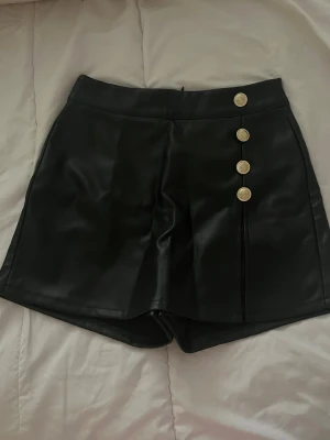 Svarta högmidjade skinnshorts - Snygga svarta högmidjade shorts i skinnimitation med fyra dekorativa guldfärgade knappar framtill. Klassisk och stilren design med en lätt A-linjeformad passform. Perfekta för att lyfta vilken outfit som helst.