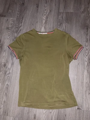 Olivgrön Moncler t-shirt med ränder - Snygg olivgrön t-shirt från Moncler med korta ärmar. Ärmsluten har coola randiga detaljer i vitt, rött och svart. Klassisk passform och mjukt bomullsmaterial. Perfekt till jeans eller shorts för en clean look.
