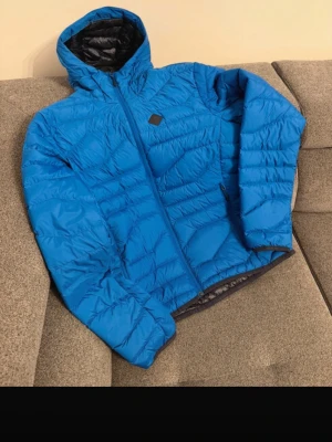 Jlindeberg blå dunjacka  - Snygg blå dunjacka med huva och dragkedja framtill. Jackan har två sidofickor med dragkedja och svart innerfoder. Perfekt för kalla vinterdagar tack vare det tjocka dunfodret. Sportig look och lätt att matcha med olika outfits.