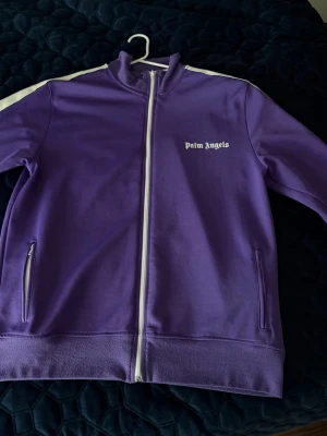 Lila Palm Angels zip hoodie - Säljer en lila hoodie från Palm Angels med vit dragkedja och vita ränder längs ärmarna. Hoodien har ståkrage, två fickor med dragkedja och logga på bröstet. Materialet är mjukt och stretchigt, perfekt för en streetwear-look.