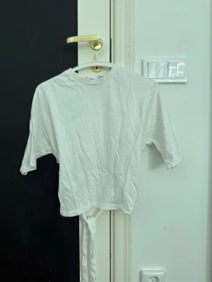 Vit croppad topp med knytband - Säljer en vit croppad topp med korta ärmar och knytband i midjan. Toppen har en enkel och clean design med rund halsringning och är perfekt för en avslappnad stil. Materialet känns som bomull och toppen har en loose passform.