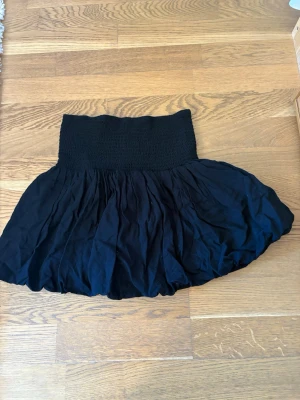 Svart kortkjol från Lindex - Svart kortkjol från Lindex med bred smock i midjan och voluminös, luftig kjoldel. Kjolen har en enkel och stilren look som passar till många olika outfits. Perfekt för dig som gillar en bekväm och trendig stil.