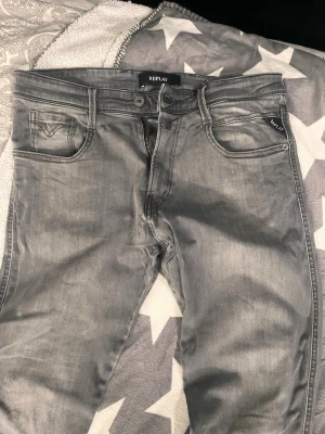 Grå jeans från Replay anbass 32/34 - Snygga grå jeans från Replay med klassisk femficksmodell och diskreta slitningar. Jeansen har dragkedjegylf och är tillverkade i bomull med lite stretch för extra komfort. Perfekta för en avslappnad och trendig look.