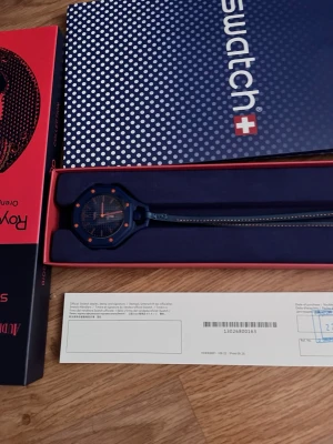 Blå Swatch x Audemars Piguet klocka - Säljer en blå Swatch x Audemars Piguet Royal Oak klocka med orange detaljer. Klockan har en åttakantig urtavla och ett matchande blått armband. Kommer i originalförpackning med tillhörande certifikat och påse. Perfekt för dig som gillar unika samarbeten!