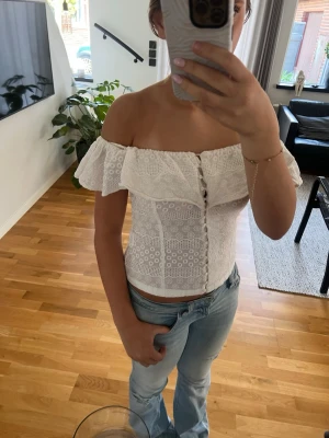 Vit offshoulder spetsblus - Säljer en superfin vit offshoulder blus med spetsdetaljer och knappar framtill. Toppen har volang runt axlarna och är figurnära med en romantisk vibe. Perfekt till jeans eller kjol för en söt look.
