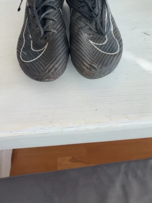 Svarta Nike Mercurial fotbollsskor - Säljer ett par svarta Nike Mercurial fotbollsskor med vita detaljer och snörning. Skorna har en modern, strömlinjeformad design och är tillverkade i syntetmaterial för bra grepp och känsla på planen. Perfekta för dig som vill ha stilrena och funktionella skor.