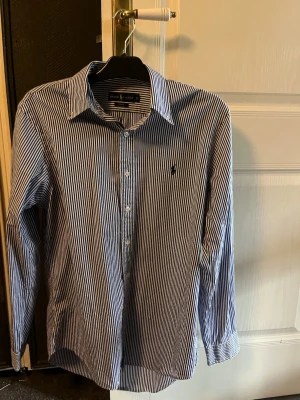 Randig skjorta från Ralph Lauren - Säljer en klassisk blåvit randig skjorta från Ralph Lauren i slim fit-modell. Skjortan har långa ärmar, knappar framtill och den ikoniska loggan broderad på bröstet. Perfekt för dig som gillar stilrena och tidlösa plagg.