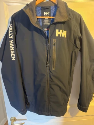 Marinblå vindjacka Helly Hansen - Snygg marinblå vindjacka från Helly Hansen med vit logga på bröstet och ärmen. Jackan har hel dragkedja, bröstficka med dragkedja och grå fleecefodrad krage. Perfekt för blåsiga dagar och outdoor-aktiviteter.