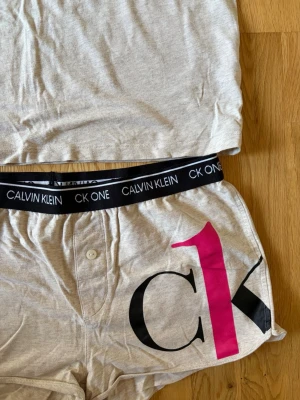 Grått CK ONE set från Calvin Klein - Snyggt grått pyjamasset från Calvin Klein med kortärmad t-shirt och shorts. Setet har svarta och rosa CK ONE-loggor, samt svart resår med vit text i midjan. Tillverkat i mjukt och stretchigt material, perfekt för chill dagar hemma.