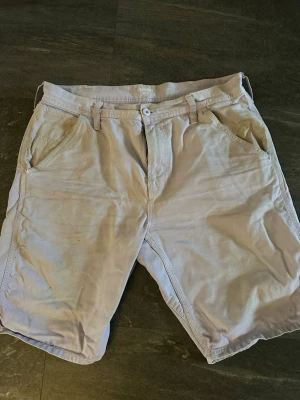Beiga shorts från Carhartt - Säljer ett par beiga shorts från Carhartt med klassisk passform. Shortsen har bälteshällor, knapp och dragkedja framtill samt två stora bakfickor. Tillverkade i slitstark bomull, perfekta för sommardagar.