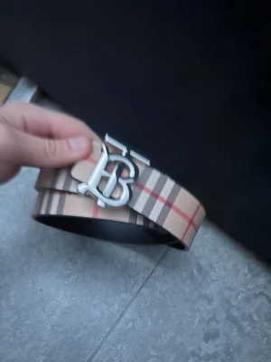 Burberry bälte  - Snyggt bälte i beige med klassiskt rutigt mönster och ett stort metallspänne med bokstäver. Bältet har svarta insidor och är tillverkat i skinn. Perfekt accessoar för att lyfta din outfit.