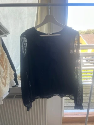Svart transparent blus med volang - Snygg svart blus från Samsøe Samsøe i transparent material med diskreta prickar och volangdetalj framtill. Blusen har långa ärmar och en luftig passform, perfekt för att lyfta vilken outfit som helst. Stor i storleken!