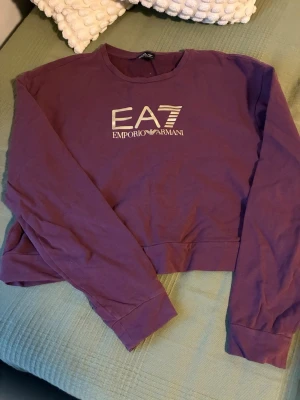 Lila cropped sweatshirt EA7 Armani - Snygg lila cropped sweatshirt från EA7 Emporio Armani med tryckt logga framtill. Tröjan har rund halsringning och långa ärmar. Perfekt för en trendig och avslappnad stil.