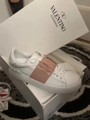 Valentino Garavani vita sneakers - Säljer ett par snygga vita sneakers från Valentino Garavani med bred ljusrosa läderdetalj över snörningen. Skorna har klassisk låg profil, rund tå och vita snören. Perfekta för dig som vill ha en stilren men unik look.