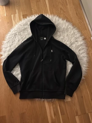 Svart hoodie från Polo Ralph Lauren - Snygg svart hoodie med dragkedja från Polo Ralph Lauren. Klassisk design med huva, dragsko och fickor framtill. Ikoniska vita loggan broderad på bröstet. Tillverkad i mjukt bomullsmaterial som är skönt att bära.