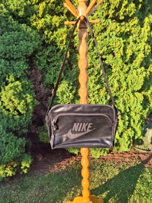 Svart axelväska från Nike - Snygg svart axelväska från Nike med stort vitt logotryck på framsidan. Väskan har justerbar axelrem, dragkedja upptill och en extra ficka framtill. Tillverkad i ett glansigt konstlädermaterial som ger en retro känsla. Perfekt för att bära det viktigaste. Kontakta gärna mig vid funderingar!