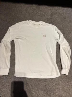 Vit långärmad tröja Calvin Klein - Vit långärmad tröja från Calvin Klein med diskret CK-logga på bröstet. Tröjan har rund halsringning och ribbade muddar vid ärmslut och nederkant. Perfekt basplagg med enkel och clean design. PRUTBART!