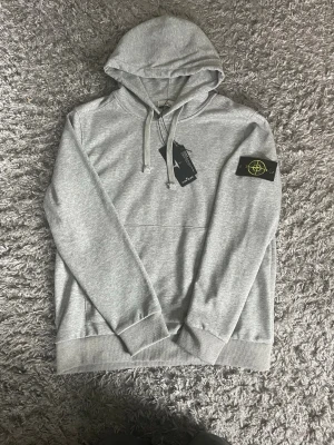 Grå hoodie med Stone Island patch - Snygg grå hoodie från Stone Island med klassisk svart och gul patch på vänster ärm. Hoodien har huva med dragsko, ribbade muddar och magficka. Tillverkad i mjuk bomullsblandning som känns skön mot huden. Perfekt för en avslappnad och trendig stil.