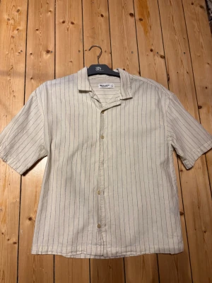 Beige skjorta med tunna ränder Pull&Bear - Kortärmad skjorta i beige med tunna vertikala svarta ränder från Pull&Bear. Skjortan har klassisk krage och knäppning framtill. Tillverkad i ett lätt tyg som passar perfekt till sommaren. Enkel och stilren design som funkar till många olika outfits. PRUTBART!